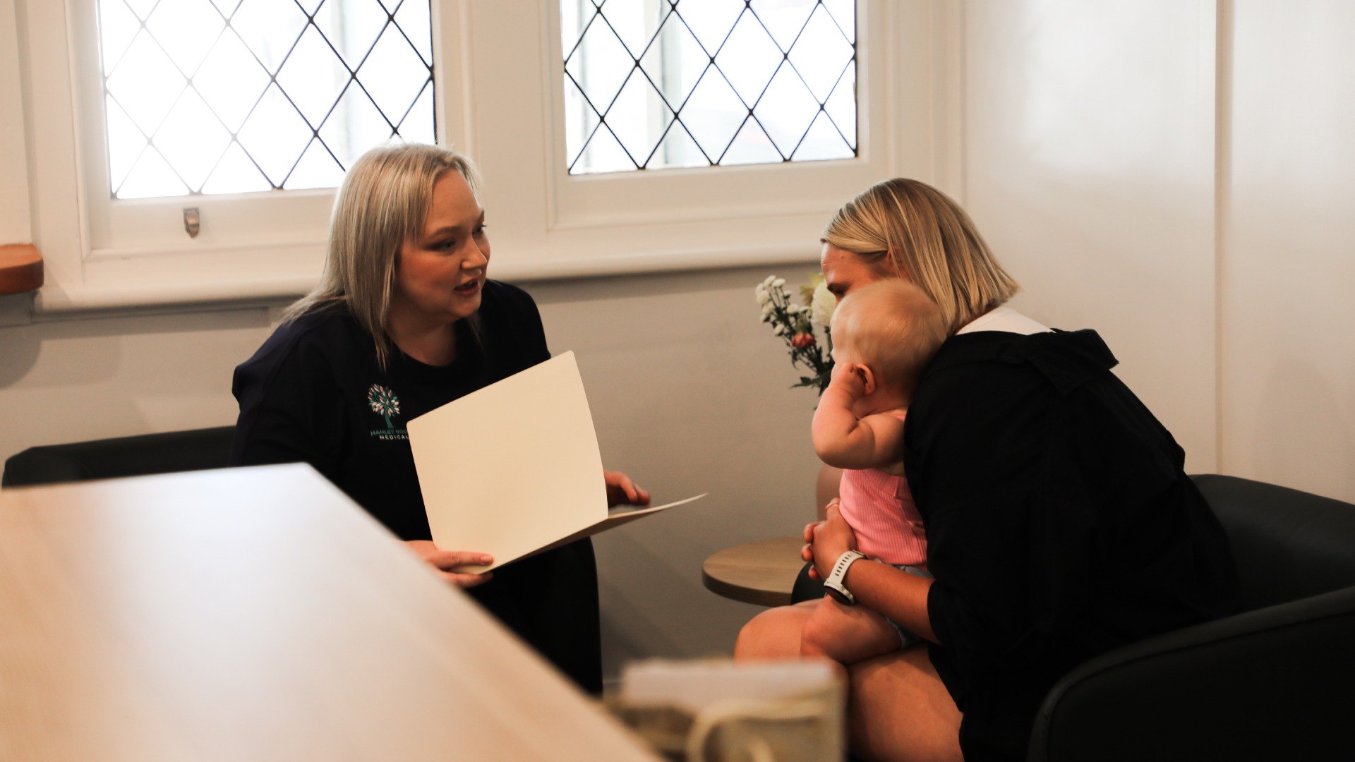 A clinician at a desk talks with a mother holding her toddler, under the clinic's leadlight windows
