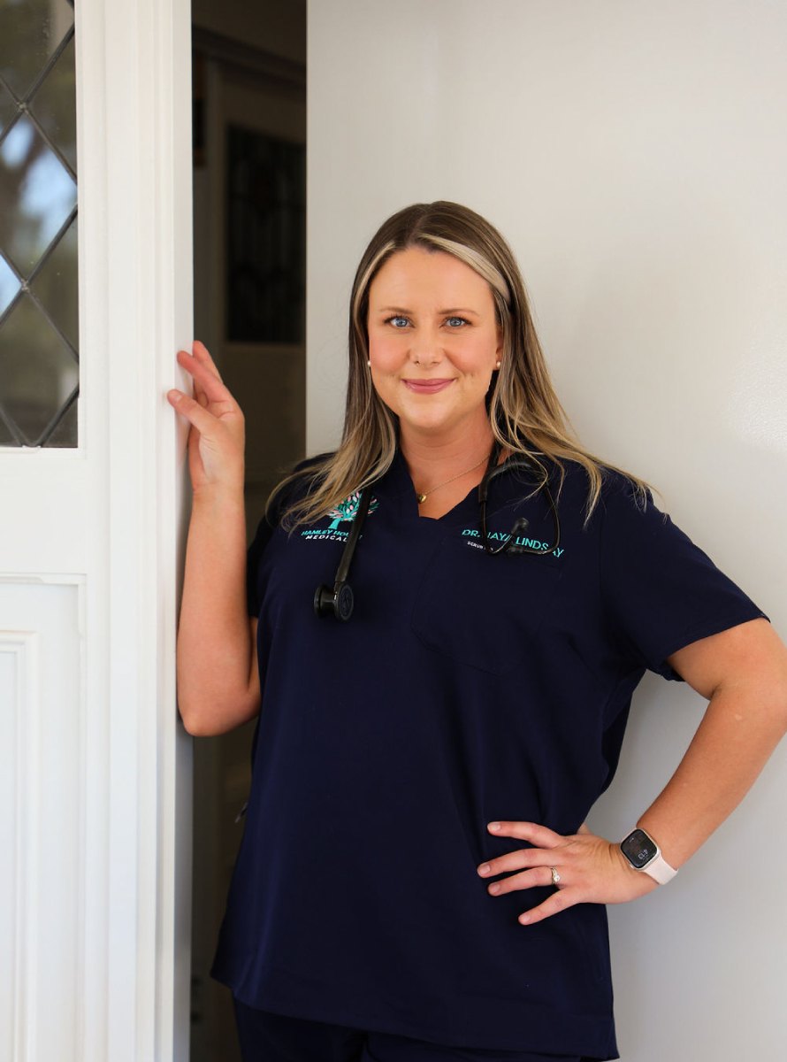 Dr Jaya Lindsay leaning against a white door frame, smiling